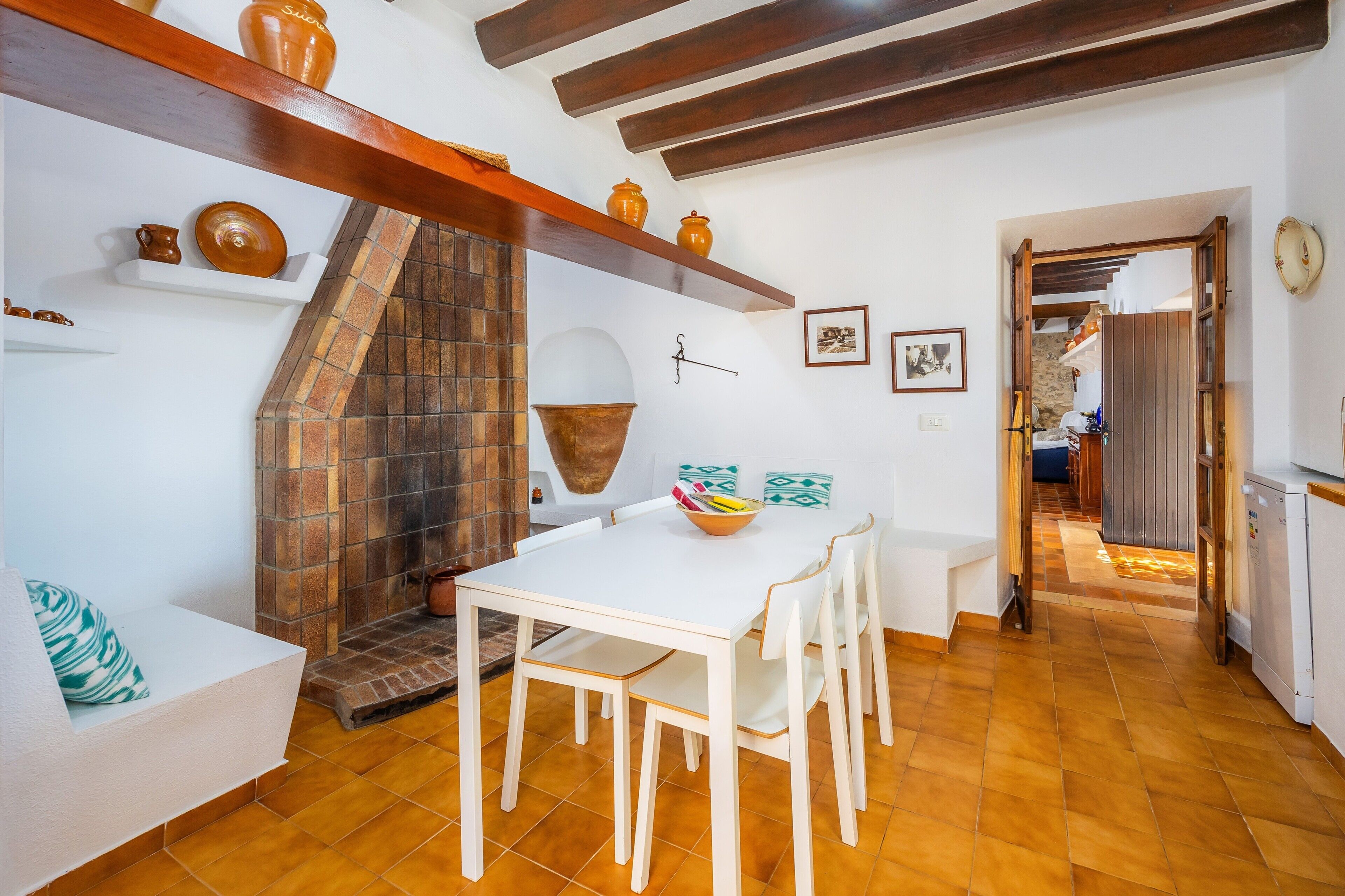 Uma sala de jantar com uma mesa e cadeiras brancas, uma lareira de tijolos e prateleiras com cerâmica.