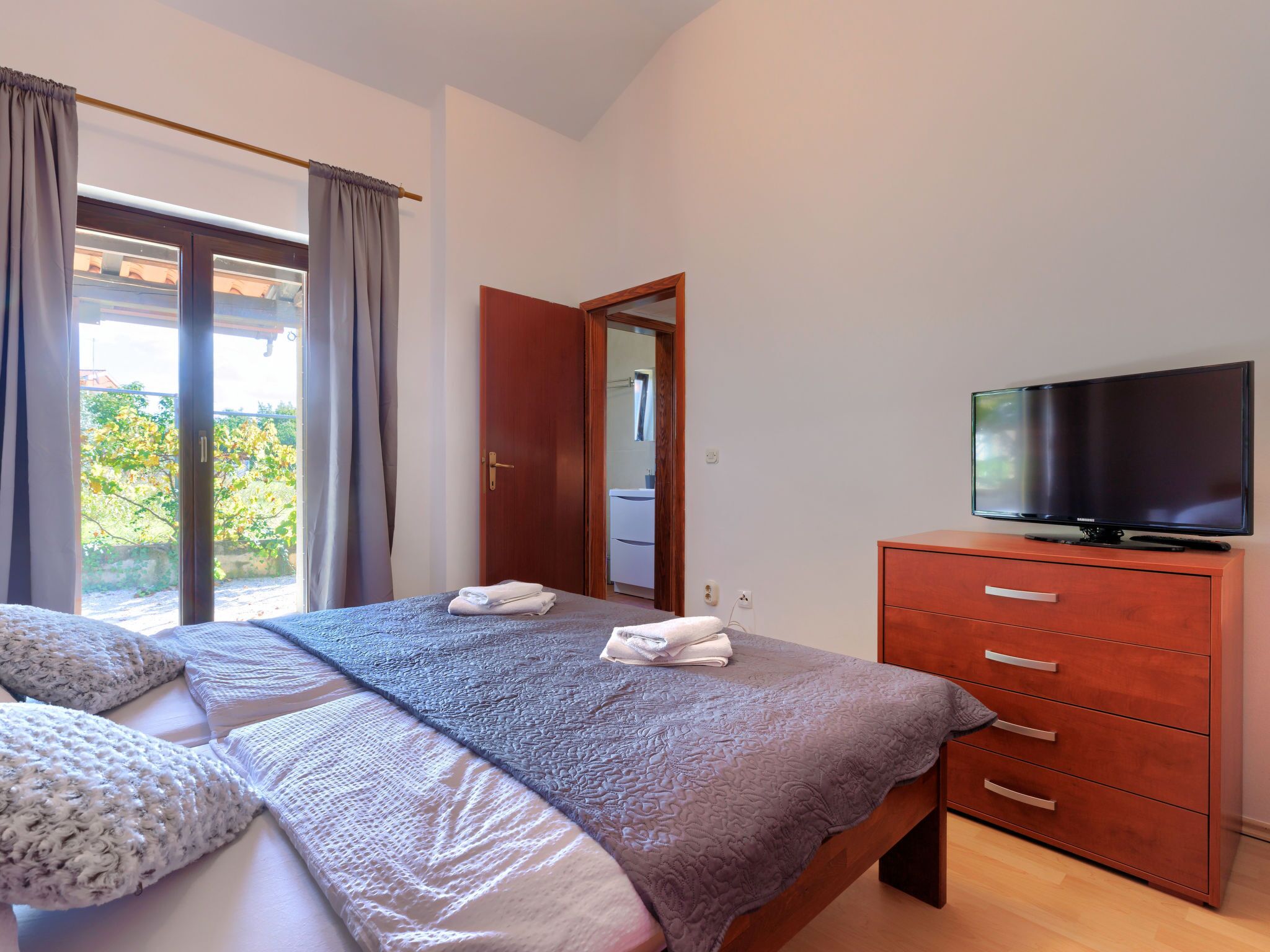 Een slaapkamer met een bed, een televisie op een dressoir en een schuifpui naar een balkon.