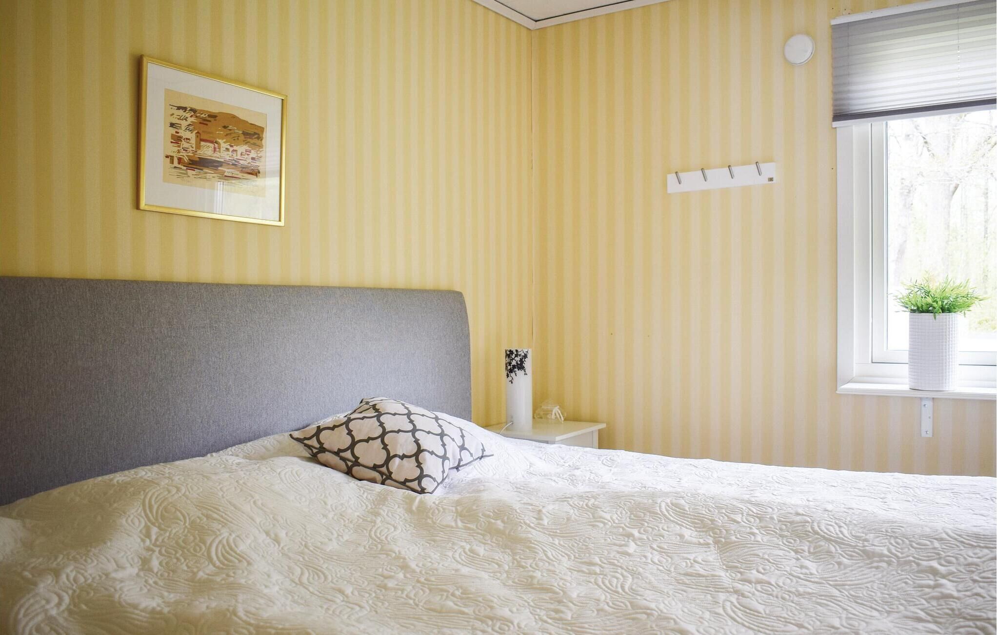 Una cama bien hecha con un cojín estampado y un cabecero, una mesita de noche con un florero, y una ventana con una planta.