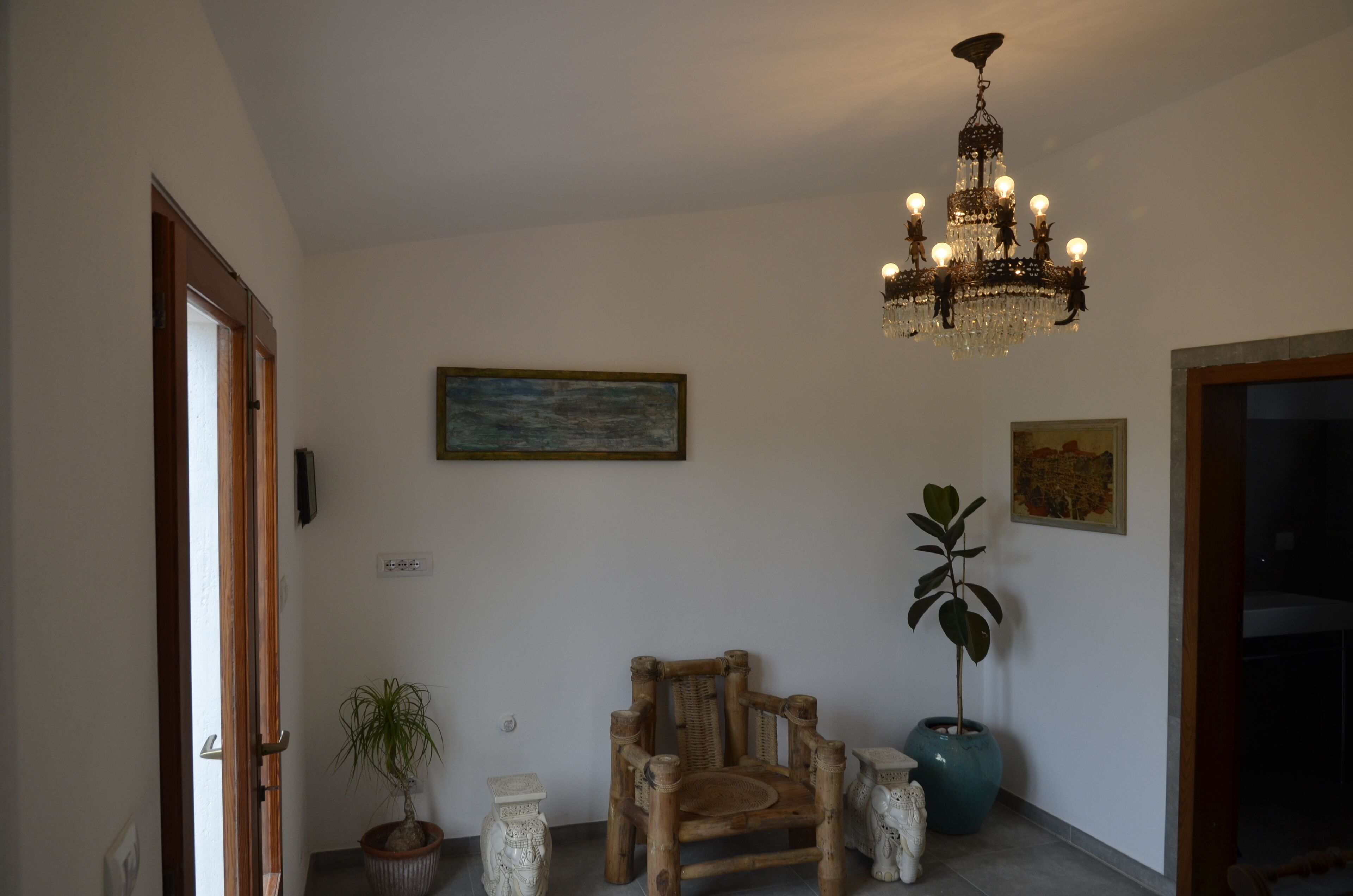 A room with a wooden chair, a potted plant, a chandelier, and two wall-mounted paintings.