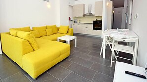 A bright yellow sofa in a modern living area with a small white table, a kitchen with white cabinets, and a dining area with a white table and chairs. - Molo Lozna by Interhome (Brač/Postira)