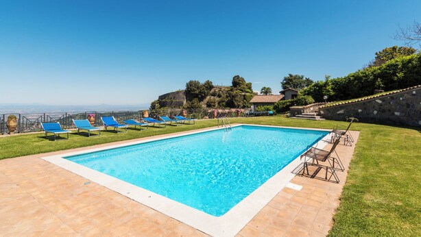 A rectangular swimming pool with blue water, surrounded by a tiled deck, and a row of blue lounge chairs.
