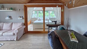Een knus interieur met een houten plafond, een bank, een tafel met een naaimachine, en uitzicht door een glazen deur op een buitenruimte.