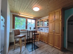 A kitchen with wooden cabinets, a dining area with two stools, a window with a view of greenery, and a ceiling with wooden beams and a light fixture.