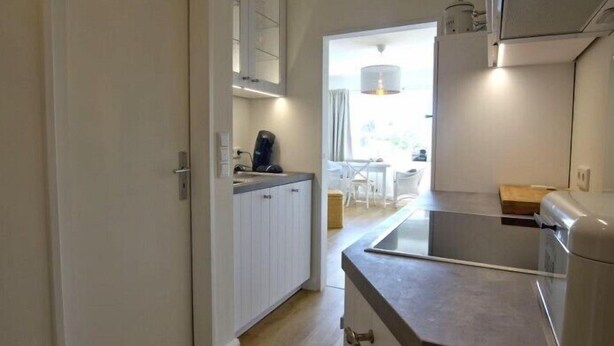 Cuina moderna amb armaris blancs, taulella i forn encastrat. Es veu una zona de menjador amb taula i cadires a través d'una porta oberta.