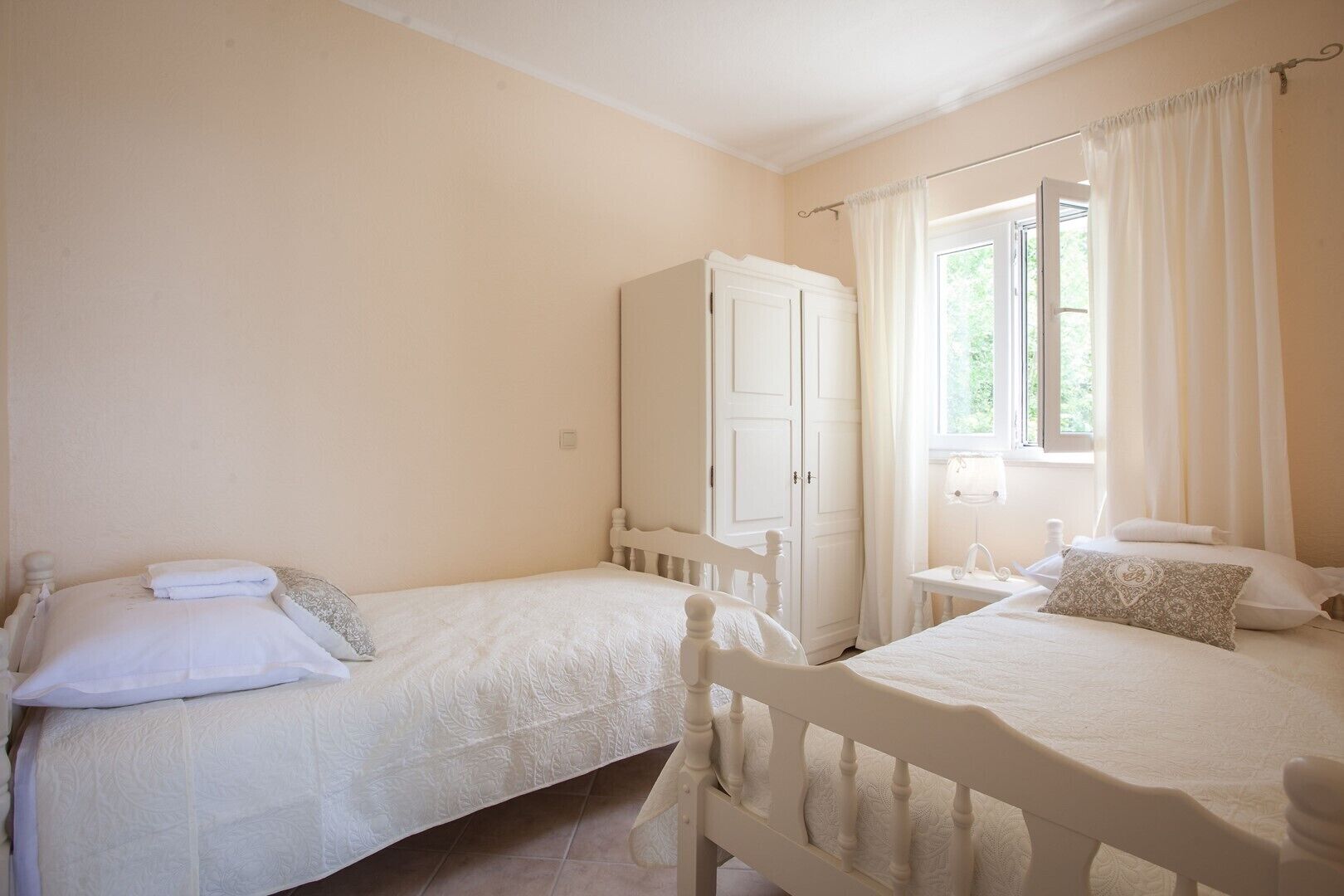 Une chambre avec deux lits simples, une armoire, une petite table de chevet avec une lampe et une fenêtre avec des rideaux.