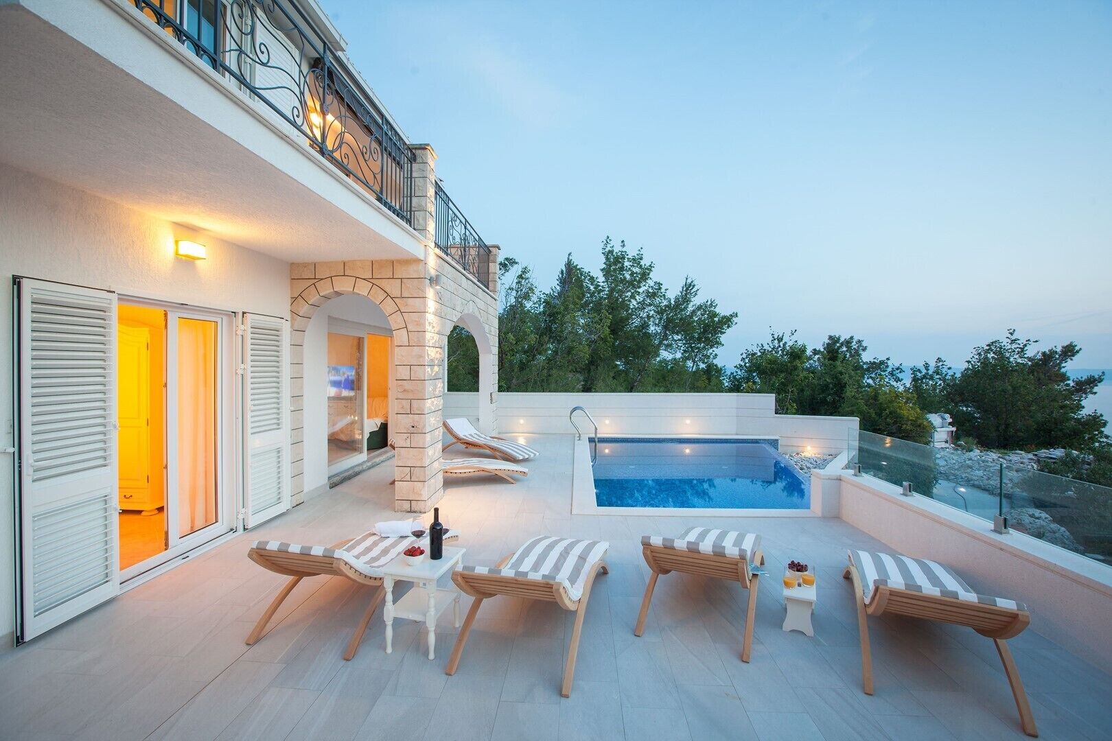 A modern outdoor pool area with lounge chairs and a hot tub, surrounded by a stone wall and overlooking a scenic landscape.