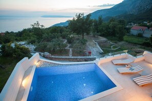 A swimming pool with a stone border and lounge chairs, surrounded by greenery and a view of the sea and mountains. - Luxury villa Retro fusion in Tucepi (Tucepi)