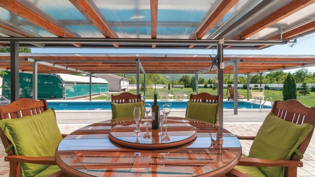 Une terrasse extérieure avec une table ronde en verre et des chaises en bois, donnant sur une piscine et un jardin aménagé.