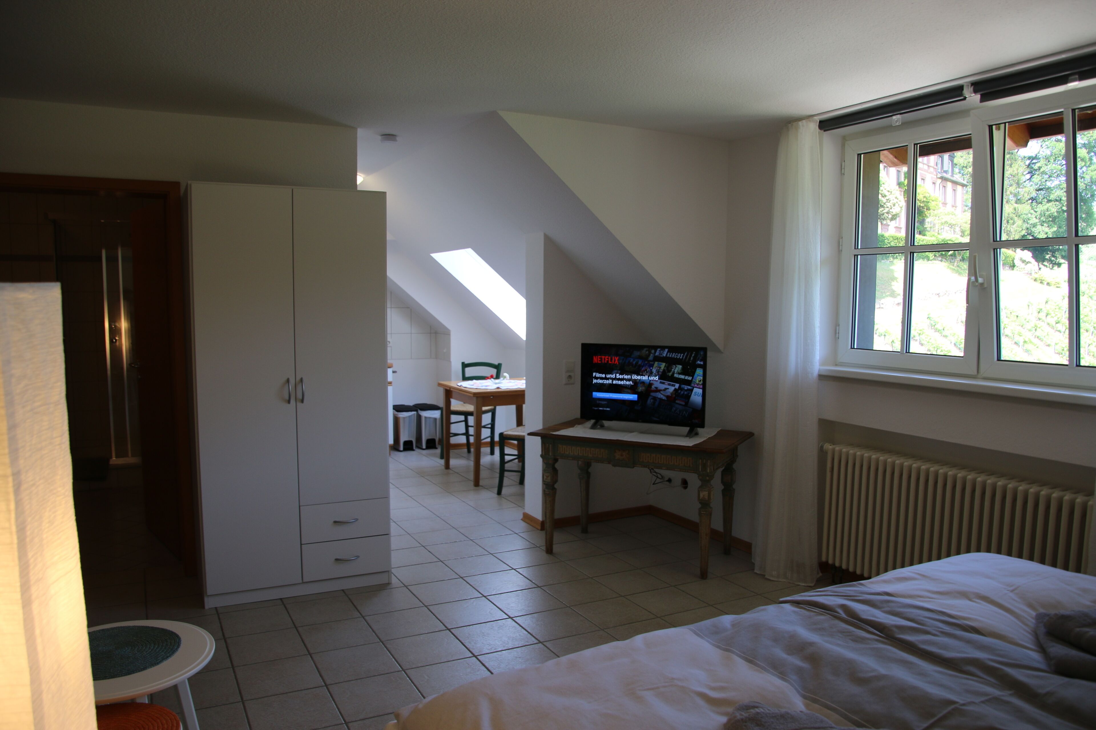A bedroom with a bed, a television, a window, and a kitchenette area.
