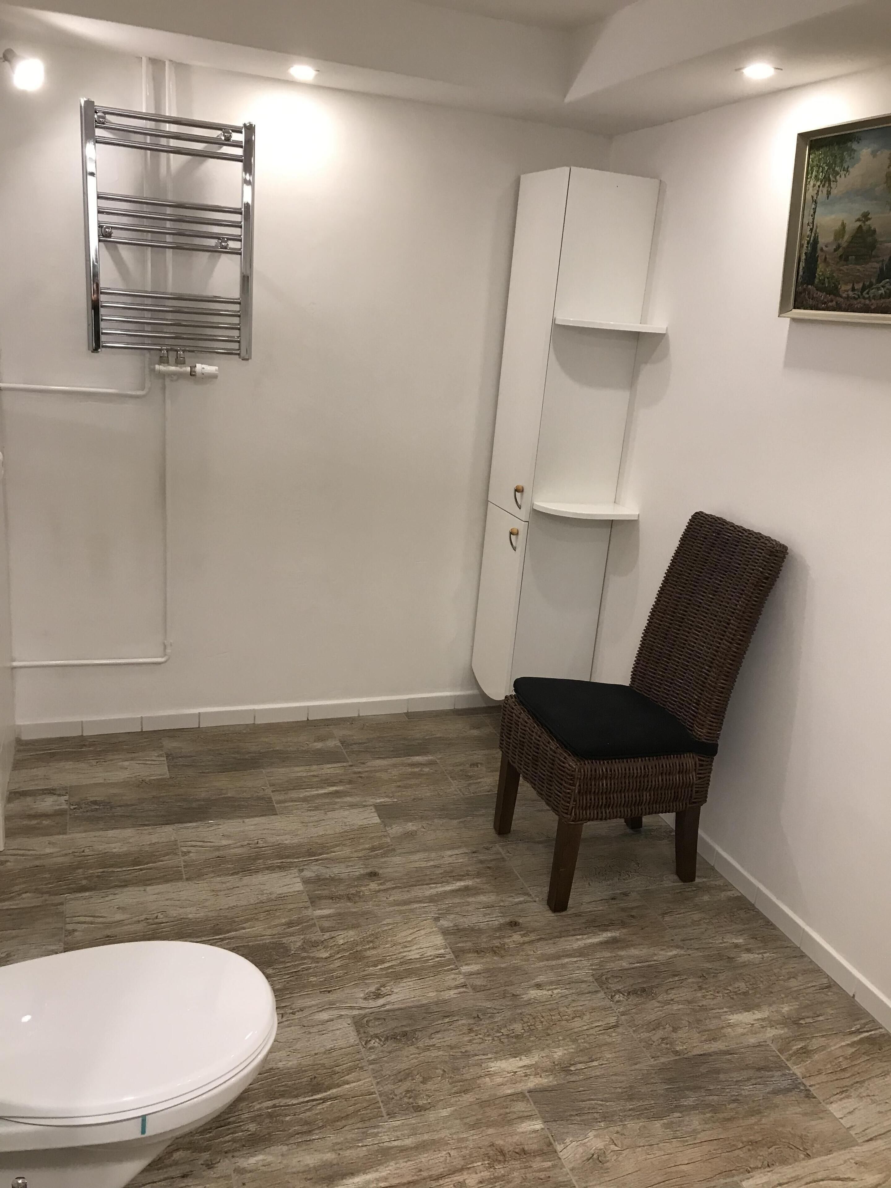 Un baño con inodoro blanco, una silla de mimbre, un armario blanco y un perchero para toallas.