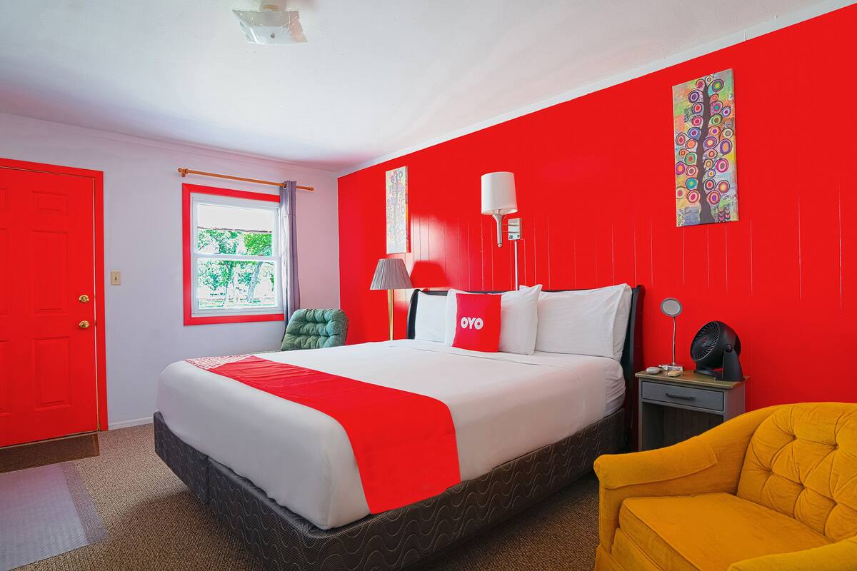 A hotel room with a red and white color scheme, a bed with a red blanket, a yellow armchair, a nightstand with a lamp, and a window with a view of greenery.