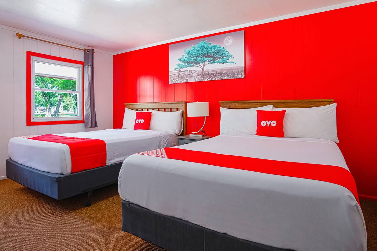 A hotel room with two beds, red and white bedding, a window with a view, and a painting of a tree on the wall.