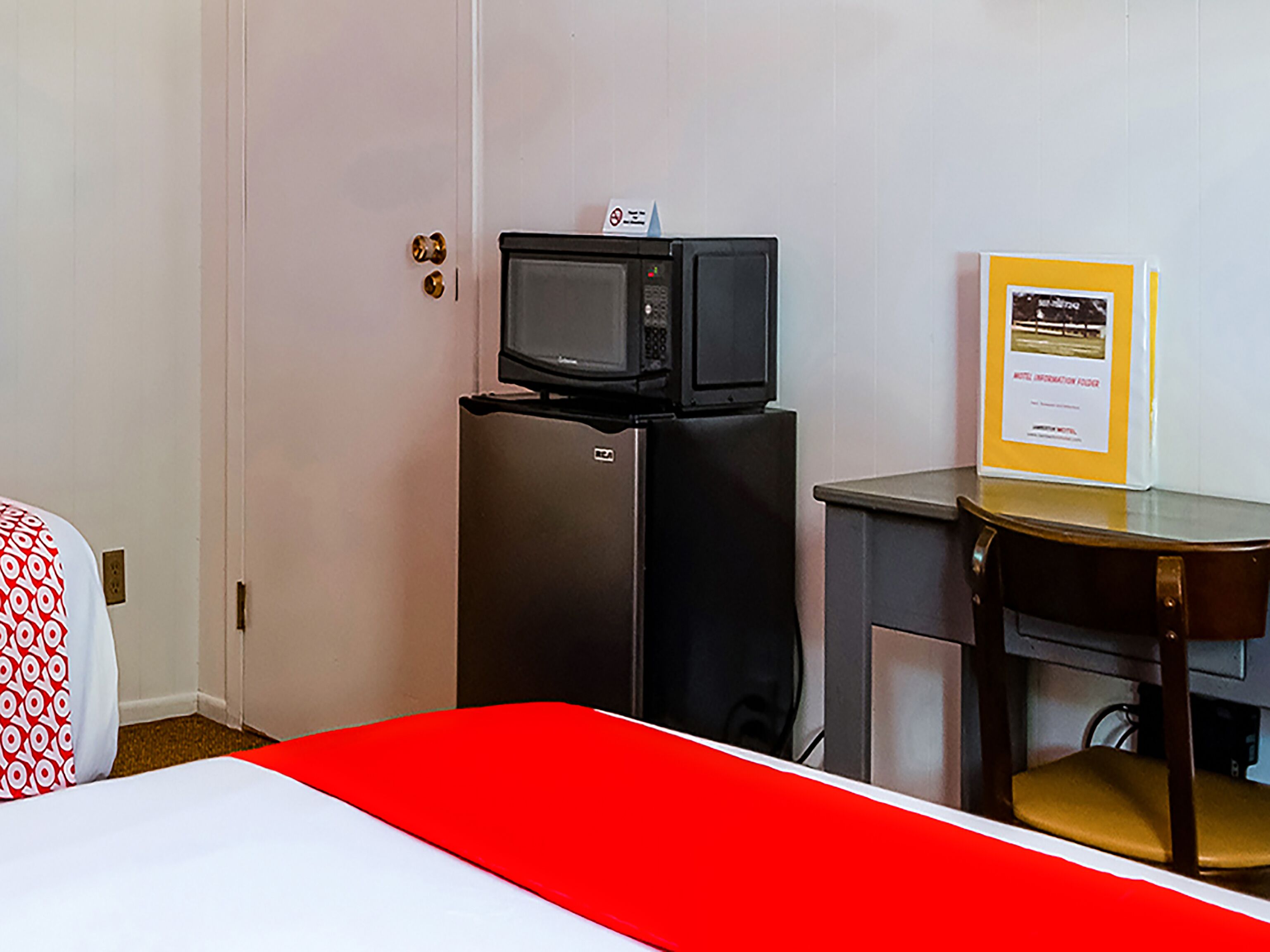 A hotel room with a bed, a small black refrigerator with a microwave on top, a desk with a chair, and a framed certificate on the wall.