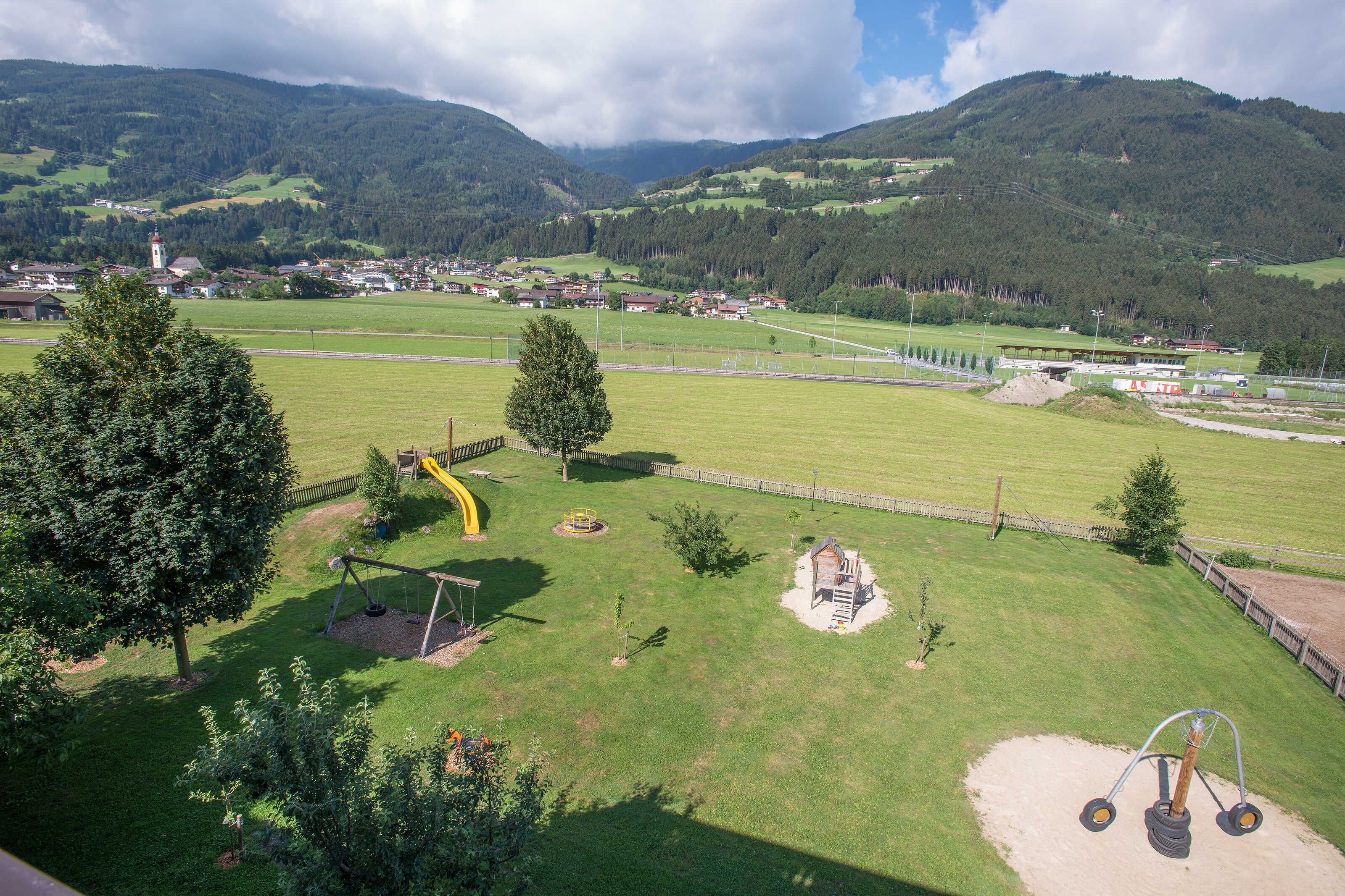 A playground with swings and a slide, surrounded by green fields and mountains in the background.