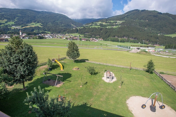 Ein Spielplatz mit Schaukeln und einer Rutsche, umgeben von grünen Feldern und Bergen im Hintergrund.