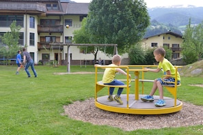 Ein Spielplatz mit Wippe und Schaukel, umgeben von einer Rasenfläche und einem Gebäude mit Balkonen.
