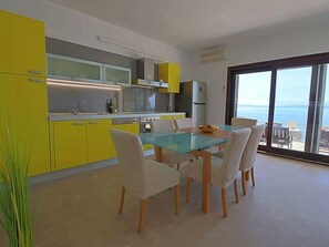 Une cuisine moderne avec des meubles de couleur jaune, une table à manger en verre et une vue sur la mer grâce à de grandes fenêtres.
