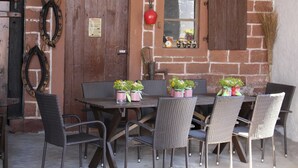 Un espace de restauration extérieur rustique, avec des chaises en osier et une table en bois, adossé à un mur de briques agrémenté d’une porte en bois et de volets aux fenêtres.