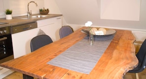 Une table de salle à manger en bois, surmontée d’une nappe grise, un vase avec une fleur blanche et une assiette contenant des petits pains.