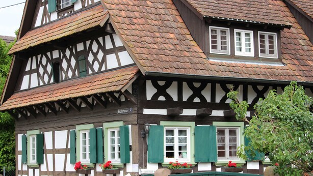 Une maison traditionnelle à colombages, dotée d’un toit à forte pente, de nombreuses fenêtres avec volets et de jardinières sur les rebords de fenêtre.