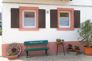 Una panchina con un cartello, piante in vaso e una ruota accanto a un edificio con finestre e persiane.