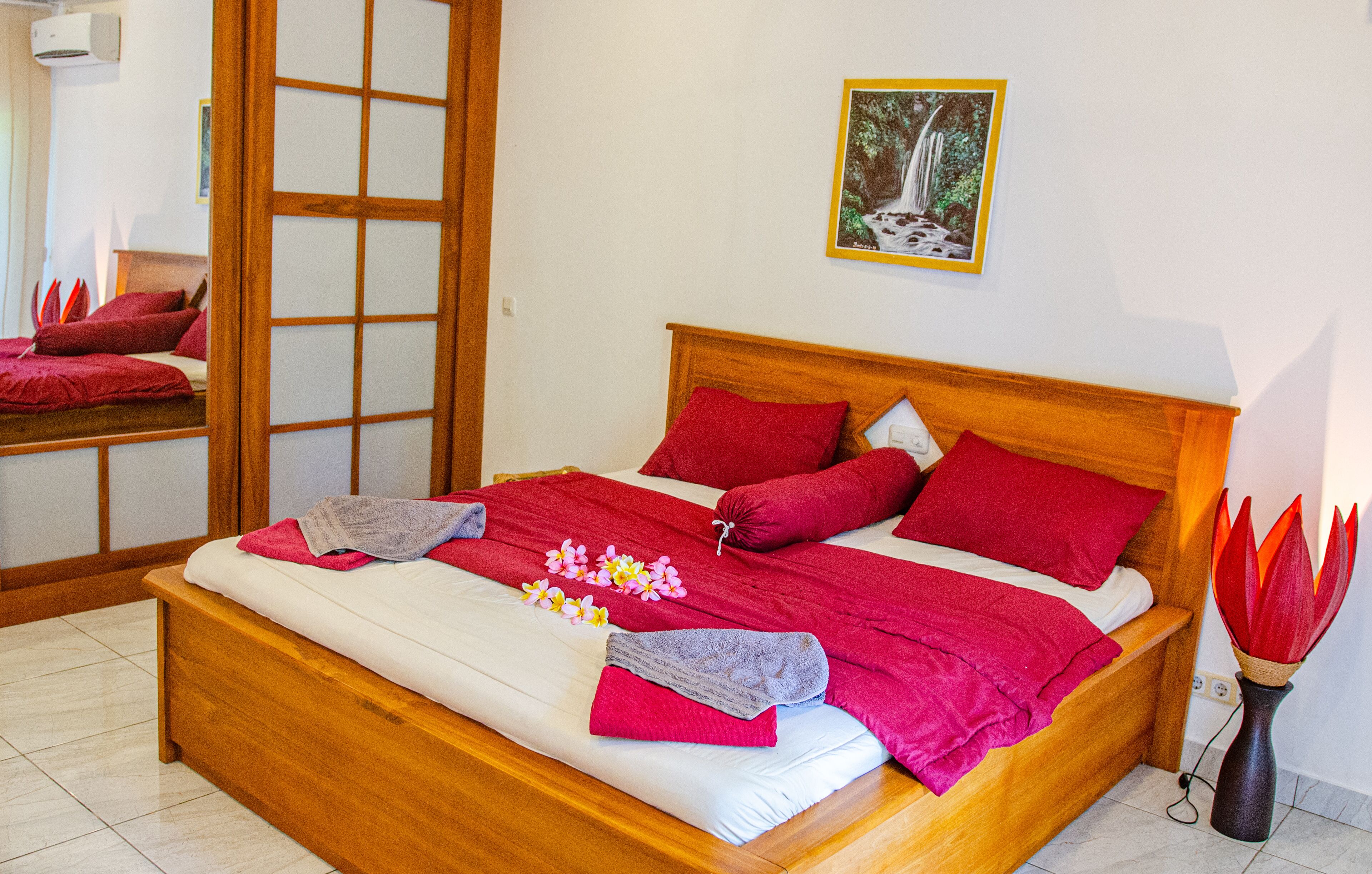 Una cama bien hecha con ropa de cama roja, un cabecero de madera y un jarrón con flores rojas a un lado.