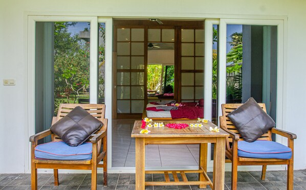 A room with two wooden chairs with blue cushions, a small wooden table with a floral arrangement, and a sliding glass door leading to a garden.