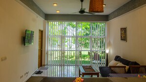 A modern living room with a large window, ceiling fan, and a flat-screen TV mounted on the wall. - Senggigi Homes & Apartments (Senggigi)