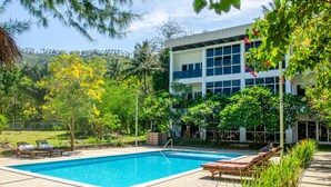 A swimming pool with lounge chairs, surrounded by lush greenery and a modern building. - Senggigi Homes & Apartments (Senggigi)