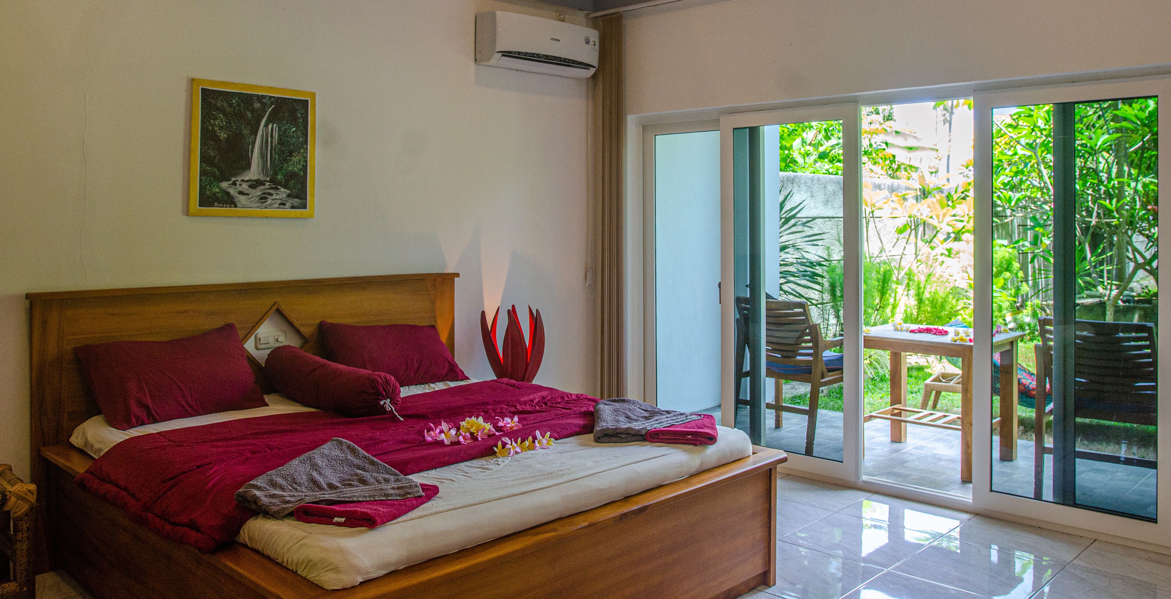 A bedroom with a wooden bed, red bedding, a painting, and a view of a patio with a table and chairs.