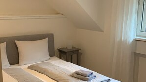 Una cama bien tendida con almohadas y toallas dobladas, una mesita de noche con una lámpara y una ventana con cortinas.