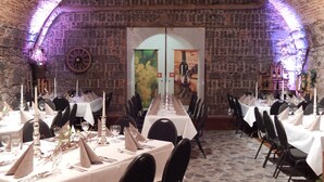 Un restaurante con paredes de piedra, mesas dispuestas con manteles blancos, sillas negras y centros de mesa con velas.