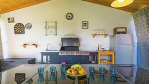 Una cocina con estufa a gas, fregadero y refrigerador. Hay vasos de vidrio azules sobre la mesa, y un frutero con manzanas y naranjas.