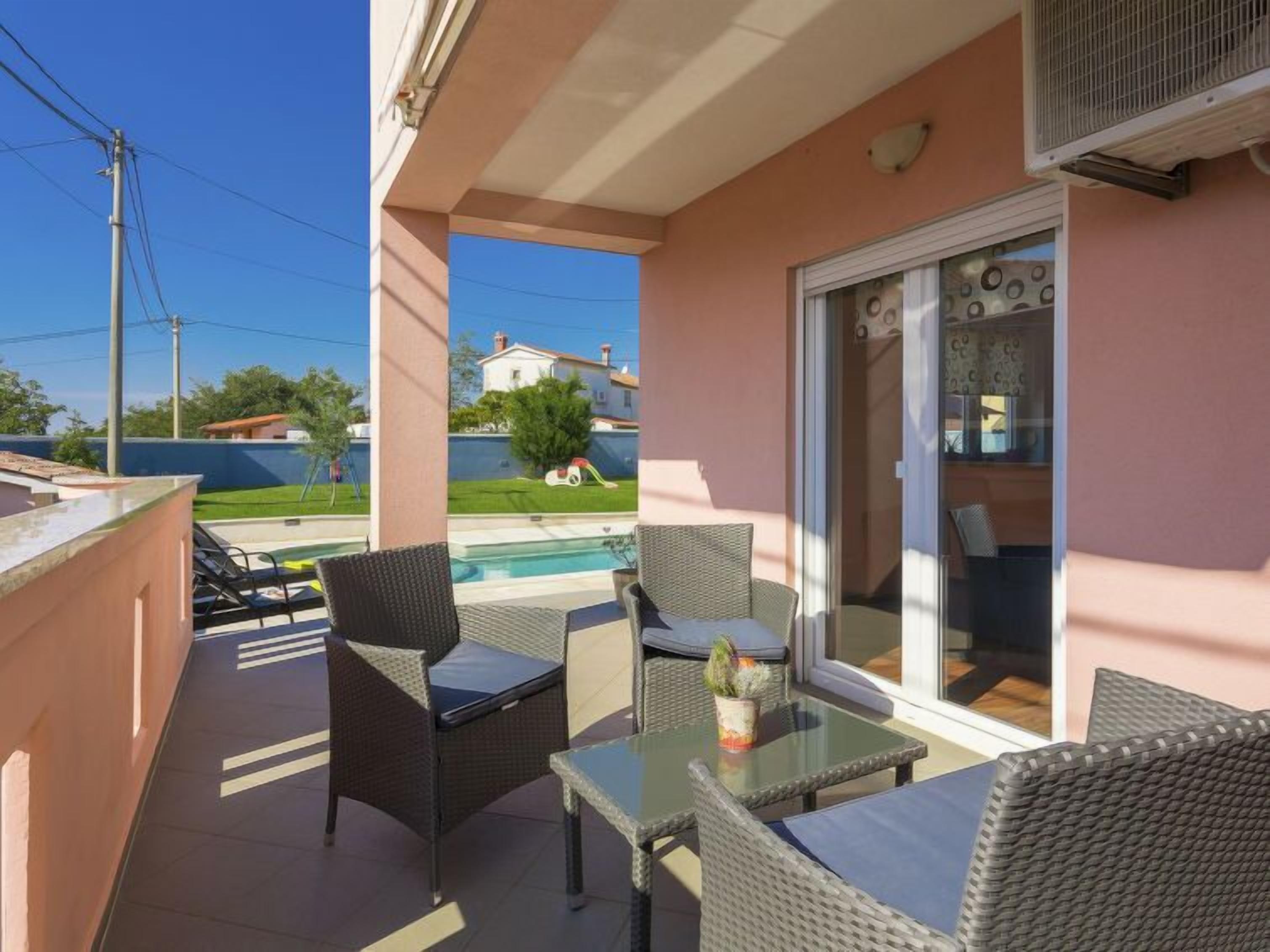 A balcony with wicker furniture, a glass table, and a view of a swimming pool and houses in the background.