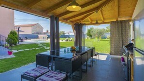 Un área de comedor al aire libre techada con mesa y sillas de vidrio, una parrilla y vistas a una piscina y jardín.