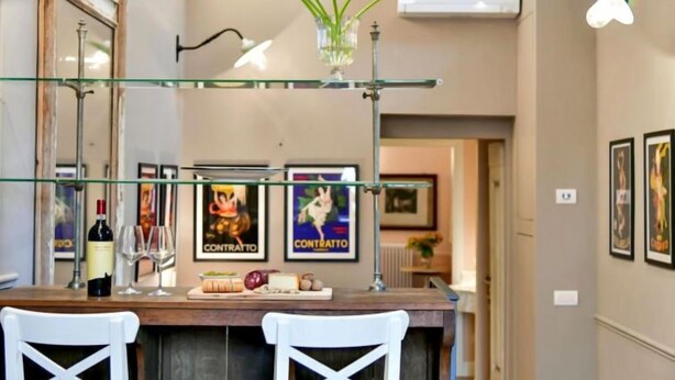 Un área de comedor con una mesa de vidrio, dos sillas blancas, un jarrón con flores y carteles enmarcados en la pared.