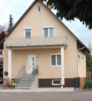 Um edifício residencial de dois andares com varanda, porta principal e escadaria que leva à entrada.
