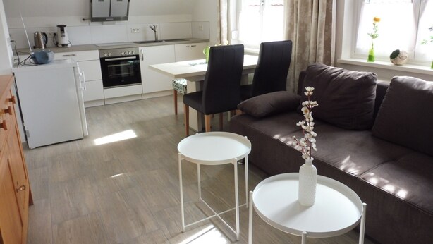 Un espacio habitable compacto con kitchenette, un sofá de cuero marrón, dos mesitas auxiliares blancas y un ventanal con cortinas.