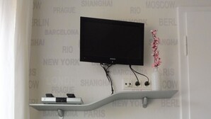 Una habitación con un escritorio blanco, una televisión montada y una estantería con dispositivos electrónicos.