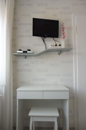 Quarto com escrivaninha branca, televisão fixada na parede e prateleira com dispositivos eletrônicos.