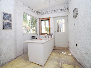 A small, white-walled room with a central island counter, a clock on the wall, and a window with a view of trees.