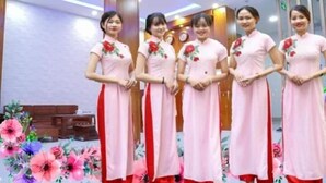 Un grupo de mujeres con vestidos rosas y detalles florales rojos posando en una habitación con muebles de madera y un arreglo floral.