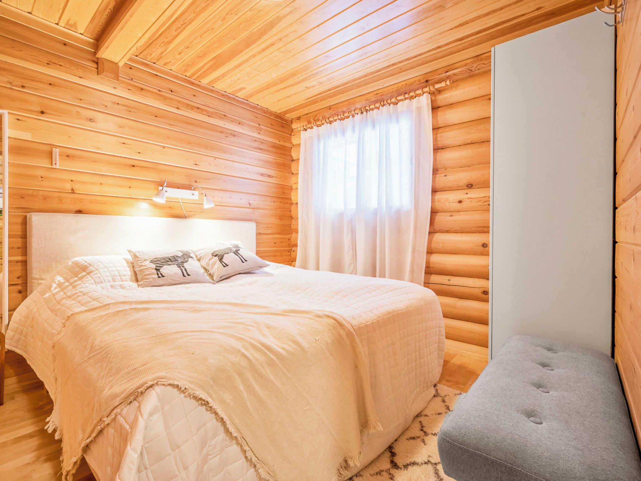 Ein Schlafzimmer einer Holzhütte mit einem Bett, Kissen, einer Nachttischlampe, einem Fenster mit Vorhängen und einer kleinen Bank.