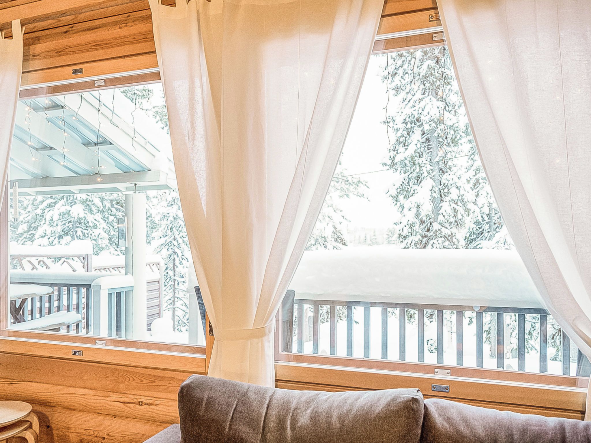 Ein Zimmer mit Holzboden, einem Fenster mit weißen Vorhängen und Blick auf ein verschneites Deck.