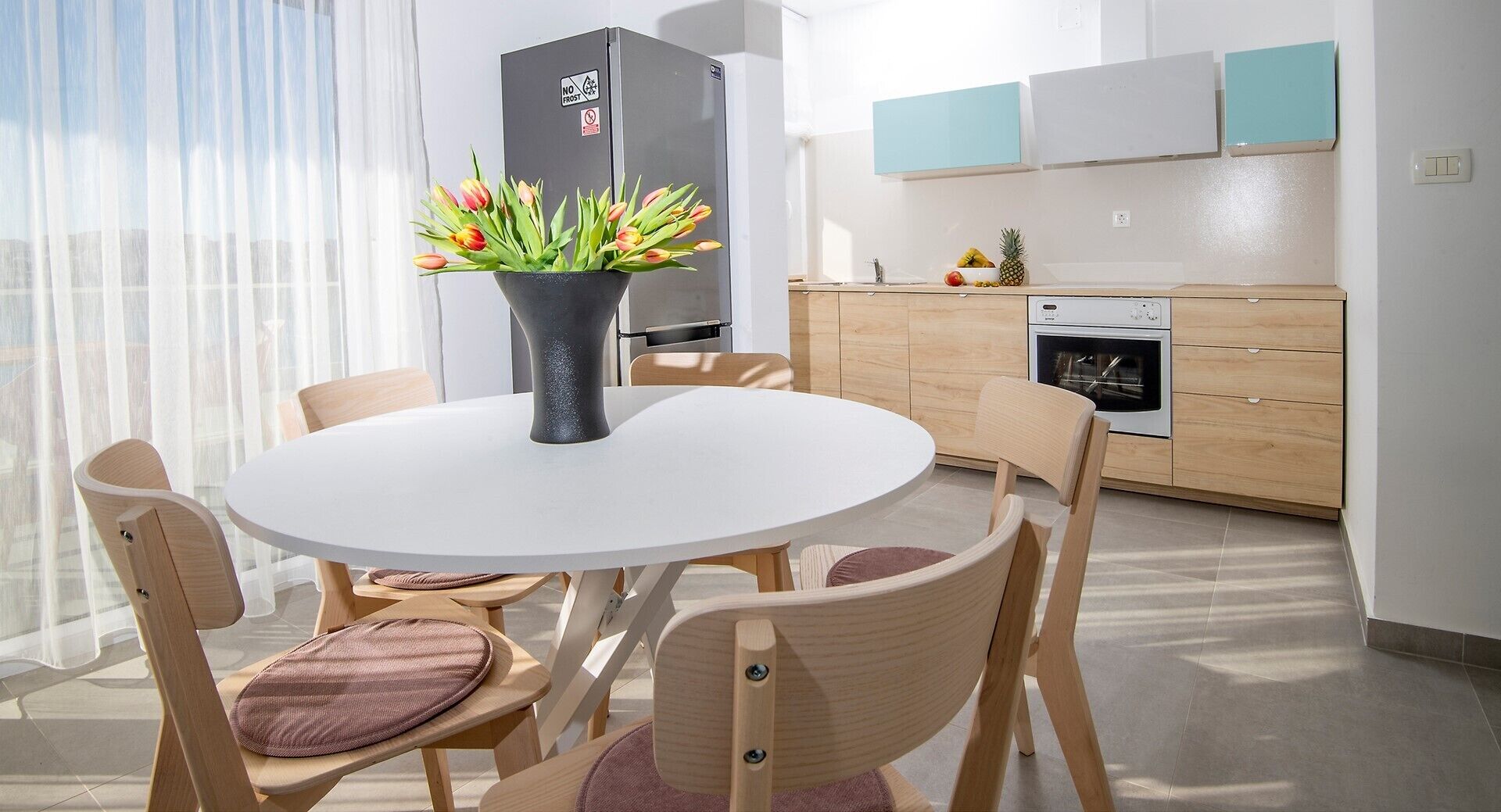 A modern kitchen with a refrigerator, oven, and a dining area with a round table and wooden chairs.