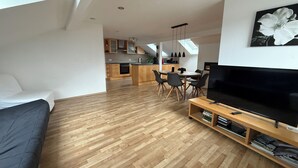 A modern living room with a large flat-screen TV, wooden flooring, a sofa, and a dining area with chairs.