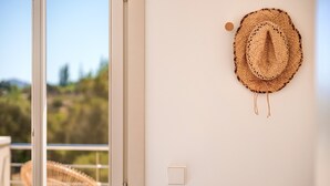 Una habitación con pared blanca, un sombrero tejido colgado en la pared, una silla de mimbre y una puerta que da a un área exterior.