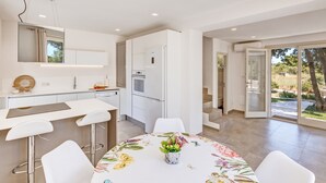 Una cocina moderna con armarios blancos, una isla central con taburetes de bar, una mesa de comedor con mantel floreado y vistas al exterior a través de amplias puertas correderas de vidrio.