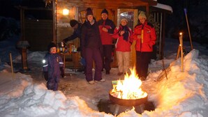 A group of people gathered around a wooden cabin with a fire in the snow.